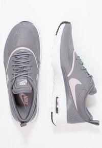 Pár šedých tenisek Nike Air Max Thea s bílými podrážkami, růžovými logy swoosh a síťovaným svrškem, zobrazených z horního a bočního pohledu na bílém pozadí.