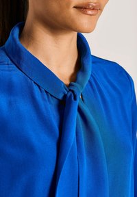 Blaue Bluse mit weichem Material, die ein gebundenes Halsdetail und eine glatte Oberfläche aufweist. Der Kragen ist gut definiert und fällt elegant.