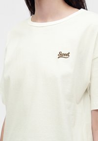 Άτομο που φοράει κρεμ T-shirt με καφέ κεντημένο κείμενο "Sweet" στο αριστερό στήθος, ορατό από τον λαιμό μέχρι τη μέση του κορμού.