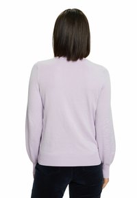 Femme aux cheveux raides et foncés portant un pull à manches longues violet clair et un pantalon sombre, de dos sur fond blanc.