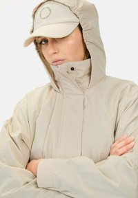 Beige wasserfeste Jacke mit Kapuze, die einen hohen Kragen und Druckknopfverschluss aufweist. Dazu gehört eine hellbeige Mütze mit einem runden Logo.