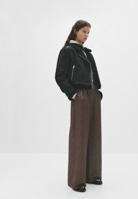 Giacca in shearling nera con design corto e chiusura con zip, abbinata a pantaloni larghi a quadri marroni e scarpe nere.