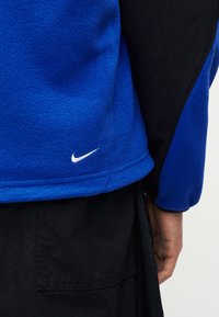 Μπλε και μαύρο φλις μπουφάν με λευκό λογότυπο Nike στο κάτω μέρος της πλάτης, φορεμένο από άτομο με ορατό δεξί χέρι και μαύρο παντελόνι.