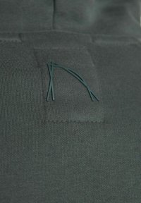 Un tessuto verde scuro presenta una superficie testurizzata con una piccola patch rettangolare che mostra un logo stilizzato in un verde complementare.