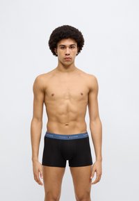 Μαύρα βαμβακερά μπόξερ με μπλε ζώνη μέσης που φέρει το λογότυπο Tommy Hilfiger. Το μοντέλο έχει μεσαία σωματική διάπλαση και σγουρά μαλλιά.