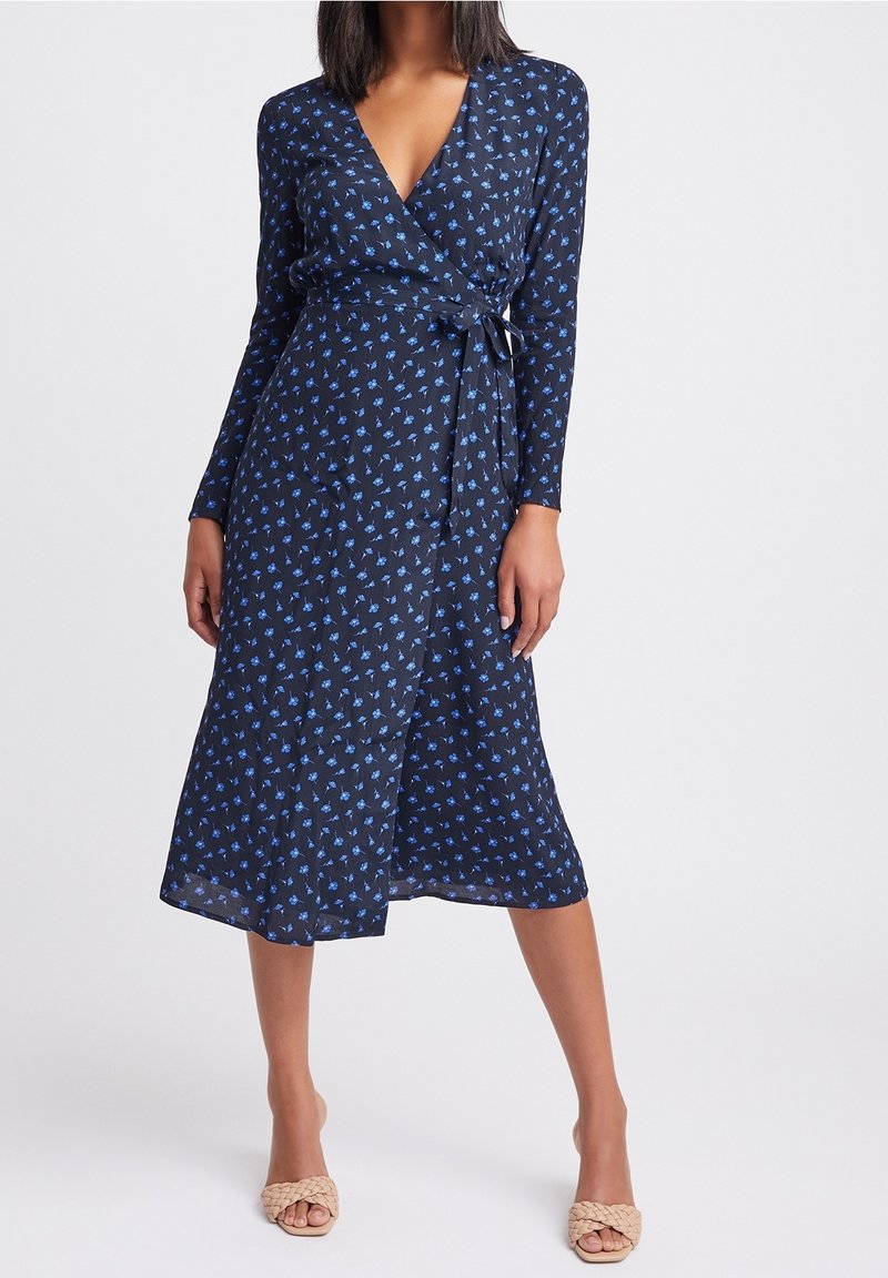 Femme portant une robe portefeuille bleu marine à imprimé floral bleu et manches longues, associée à des talons ouverts tissés beige, debout devant un fond blanc.