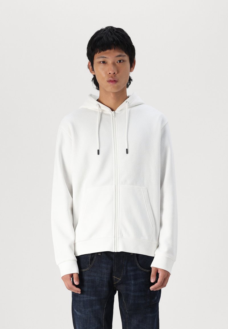 Un sweat à capuche zippé blanc avec une texture douce, doté d'une poche avant, d'une capuche avec cordon de serrage et de poignets côtelés. Porté sur un jean foncé.