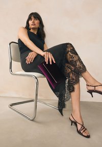 Robe noire avec ourlet en dentelle, pochette en velours violet et talons brun à brides, assise sur une chaise chromée sur un fond neutre.