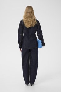 Denimjakke og bukser i mørkeblå med gul syning; modellen holder en lyseblå blød accessory. Glat tekstur og skræddersyet pasform.