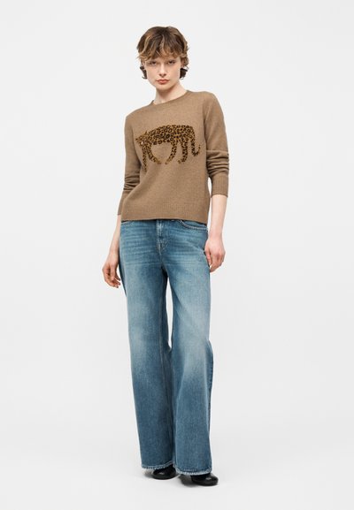 Personne portant un pull marron avec un motif léopard et un jean bleu à jambes larges, debout devant un fond clair uni.