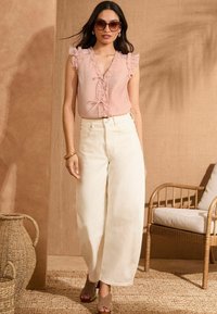 Blusa senza maniche rosa chiaro con rouches e dettaglio a fiocco, abbinata a jeans bianchi a vita alta e a gamba larga. Sandali con tacco marroni e occhiali da sole.