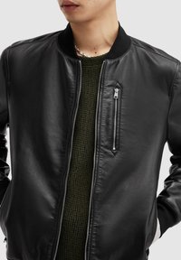 Svart läderbomberjacka med ribbad krage, blixtlåsfickor på sidan och slät yta; bärs över en mörkgrönstickad tröja.