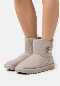 Bottes de cheville en suède gris avec un bout arrondi, des détails cousus et un accent de bouton décoratif sur le côté. Semelle extérieure plate et texturée.