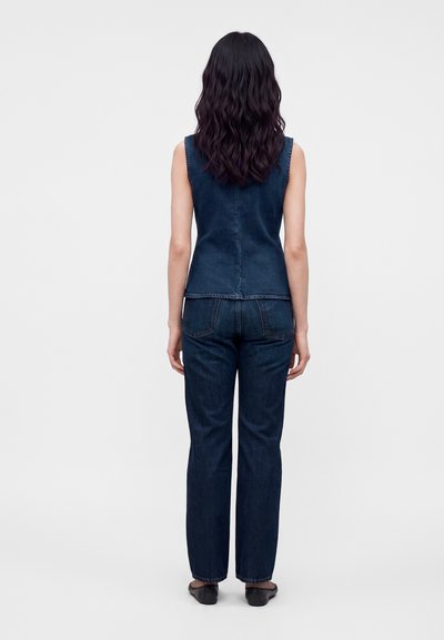 Femme aux longs cheveux ondulés foncés, vue de dos, portant un haut en denim foncé sans manches, un jean assorti et des ballerines noires pointues sur fond blanc.