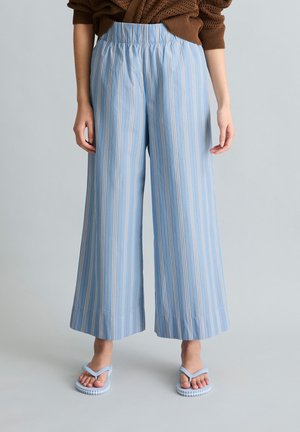 Frau trägt weit geschnittene, himmelblaue Hose mit weißen und beigen vertikalen Streifen, kombiniert mit hellblauen Sandalen und einem braunen Pullover.