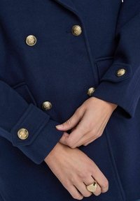 Manteau bleu marine croisé avec boutons en crête dorés, en mélange de laine texturé et avec poches latérales, au design ajusté et aux poignets retroussés.