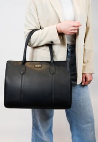 Zwarte leren tote bag met twee handvatten, voorzien van een soepele textuur, subtiele stiksels en een klein metalen logo-plaatje aan de voorkant.