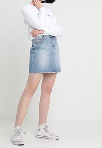 Denimminikjol i ljusblått med fransad fåll, kombinerad med en vit långärmad skjorta och vita högstövlade sneakers.