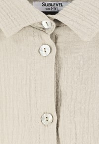 Light beige button-up shirt made of textured fabric, featuring a classic collar and three pearly buttons down the front. Size M/L.