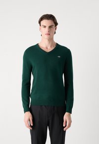 Pulover verde închis cu decolteu în V, realizat dintr-un material moale, având mâneci lungi și un mic accent cu logo pe piept.