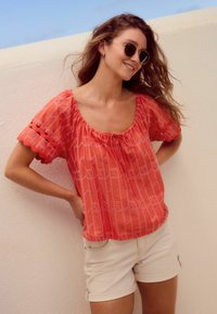 Koraalroze top met korte mouwen, een gerimpelde halslijn en geborduurde details. Gecombineerd met lichtbeige shorts, tegen een lichte achtergrond.