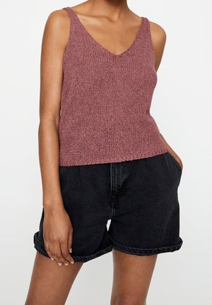 Kobieta w bezrękawniku w kolorze mauve oraz wysokich, czarnych, dżinsowych szortach z podwiniętymi brzegami, z jedną ręką w kieszeni.