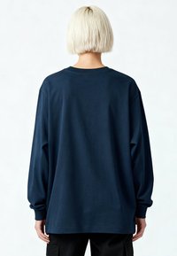 Personne aux cheveux blonds courts vue de dos, portant une chemise bleu foncé à manches longues oversize et un pantalon noir, sur un fond blanc.