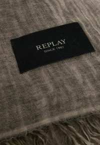 Tessuto grigio intrecciato con un sottile motivo a spina di pesce e bordi sfrangiati. Presenta un'etichetta nera con testo argentato: "REPLAY SINCE 1981."