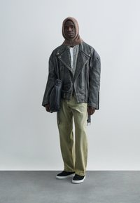 Veste de moto en cuir noir, coupe oversize, associée à une chemise à carreaux et un jean vert ample. Capuche en maille marron et baskets noires.