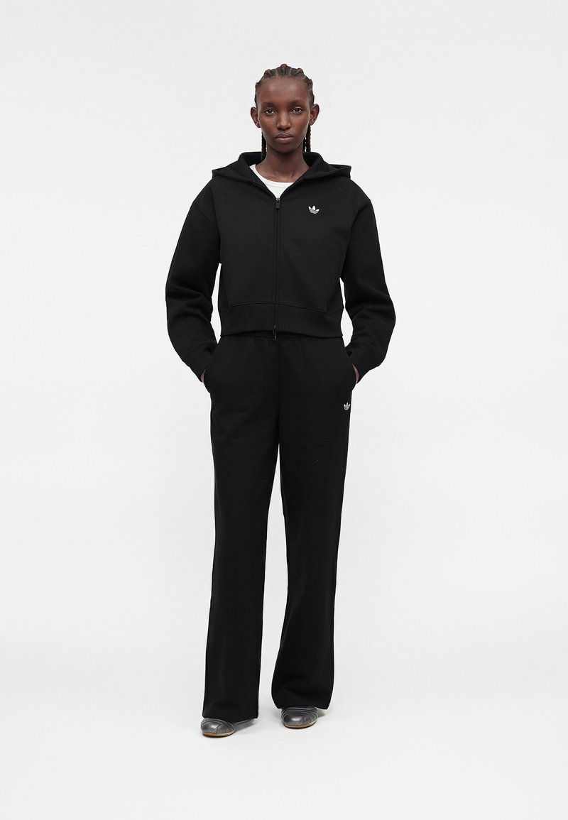 Femme aux cheveux tressés portant un sweat à capuche noir zippé et un pantalon noir assorti avec un logo blanc, debout les mains dans les poches sur fond blanc.
