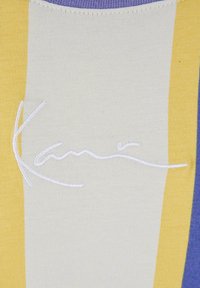 Pletený top s vertikálními pruhy v žluté, krémové a levandulové barvě, s bílým vyšívaným podpisem. Hladká textura a přiléhavý střih.