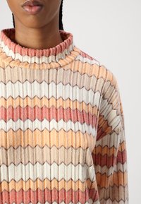 Strikket rullekragesweater med zigzag mønstre i oransje, brunt og hvitt. Har ribbestrikket krage og teksturert stoff.