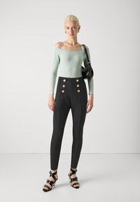 Pantaloni neri a vita alta con bottoni dorati, abbinati a un top verde menta off-the-shoulder. Il modello tiene una piccola borsa nera e indossa sandali con tacco.