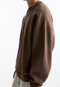 Felpa marrone con una vestibilità comoda, colletto e polsini a costine. Tessuto liscio, dettagli di cucitura minimi, abbinata a pantaloni in denim.