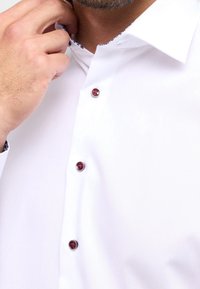 Biała koszula z klasycznym kołnierzykiem, z kontrastowym wzorzystym materiałem wewnątrz kołnierzyka i czerwonymi guzikami jako akcentami. Gładka tekstura.