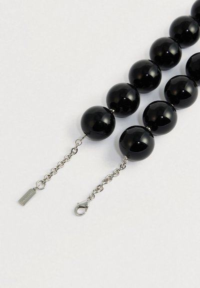 Spīdīga melna pērlīšu kaklarota ar sudraba ķēdi un vēžu aizdari uz gaiša fona.