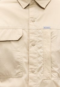 Μπεζ σακάκι με κουμπιά, με τσέπη με καπάκι στα αριστερά, ραμμένα ραφές και μια μικρή ετικέτα με το λογότυπο της Columbia στη δεξιά πλευρά του στήθους.