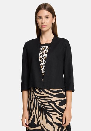 Schwarze, verkürzte Jacke mit rundem Ausschnitt und Dreiviertelärmeln, getragen über einem gemusterten Oberteil und einem Rock mit Zebra-Print.
