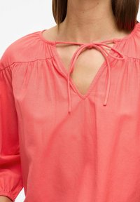 Koralfarvet bluse med V-udskæring, bindebånd ved halsen og samlede detaljer ved skuldrene. Lavet af letvægtsstof.