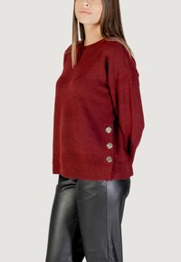 Maglione borgogna lavorato a maglia con orlo a coste e tre bottoni decorativi in argento sul lato. Abbinato a pantaloni in pelle nera.