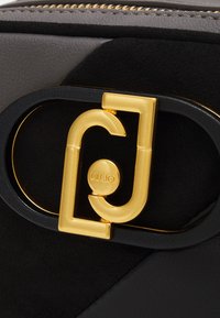 Cuir texturé noir et gris avec un fermoir en métal doré arborant un logo stylisé. Fini lisse avec des matériaux contrastants.