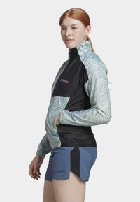 Leichte, mehrfarbige Sportjacke mit geometrischen Mustern, schwarzen und mintgrünen Bereichen, hohem Kragen und Reißverschluss. Kombiniert mit blauen Shorts.