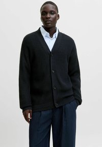 Cardigan noir en tricot avec un col en V et cinq boutons sur le devant, porté sur une chemise bleu clair à col et associé à un pantalon à rayures.