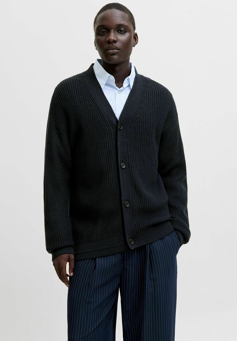 Cardigan noir en tricot avec un col en V et cinq boutons sur le devant, porté sur une chemise bleu clair à col et associé à un pantalon à rayures.