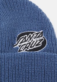 Beanie tricotat albastru cu un logo brodat alb-negru pe manșeta pliată. Materialul are un aspect texturat, cu băț.