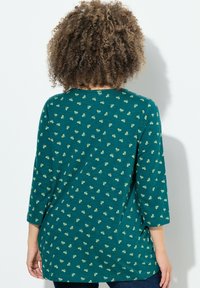 Teal langzijdige shirt met een ronde halslijn. Heeft een gestructureerde stof met kleine, verspreide lichtgroene motieven. Losse fit en gebogen zoom.