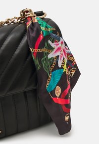 Sac à main en cuir noir avec un design matelassé. Un foulard coloré et à motifs avec des motifs floraux et en chaîne est noué à la quincaillerie dorée.