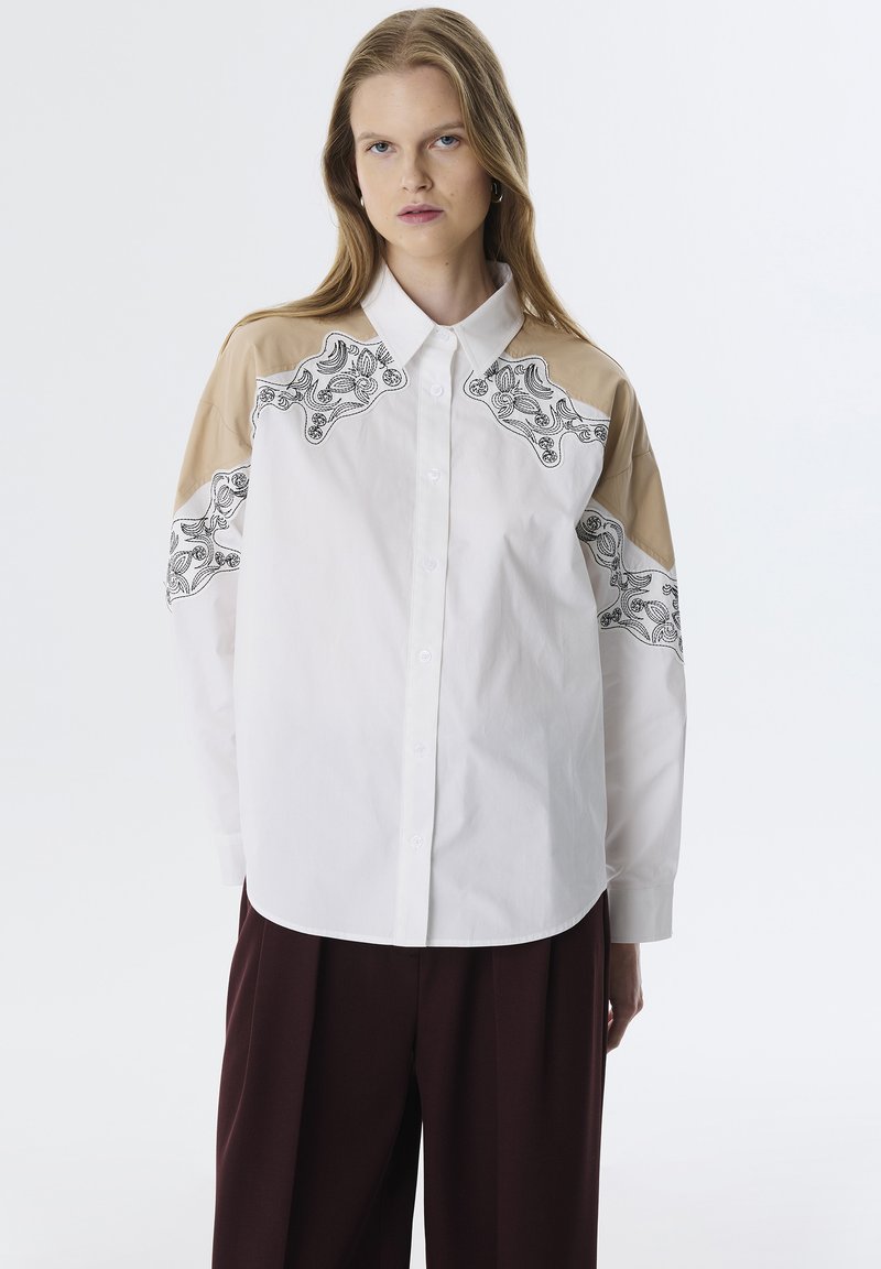 Camicia bianca con bottoni e spalle beige, caratterizzata da un elaborato ricamo floreale nero sul petto e sulle maniche. Tessuto morbido, colletto classico.