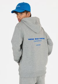 Grijze hoodie met een ruime capuchon, voorzien van "NEW BAD HABITS" in blauwe tekst op de achterkant. Gepaarde met een blauwe pet met het label "LES'ARCS."