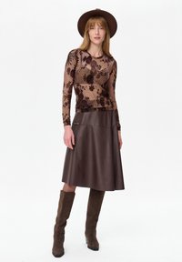 Vrouw met een brede bruine hoed, doorschijnende lange mouwen met bloemenprint, donkerbruine leren rok en kniehoge suède laarzen, staand tegen een witte achtergrond.